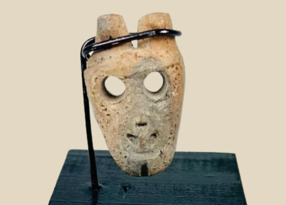 Miniature anthropomorphic mask pendant carved from bone, featuring hollowed eyes, prominent nose ridge, open mouth, and twin suspension holes; mounted on custom display stand; Western Mexico, Colima/Nayarit region, circa 300 BCE–300 CE.