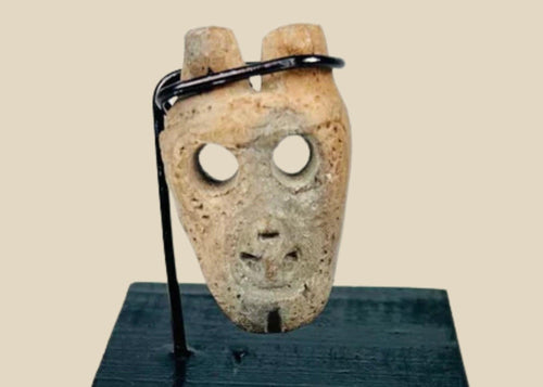 Miniature anthropomorphic mask pendant carved from bone, featuring hollowed eyes, prominent nose ridge, open mouth, and twin suspension holes; mounted on custom display stand; Western Mexico, Colima/Nayarit region, circa 300 BCE–300 CE.