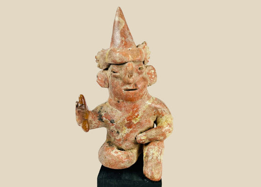 Hollow West Mexican Nayarit seated terracotta figure with tall conical headdress, turban band, large earspools, and raised right hand holding an attribute, featuring red and cream slip remnants, mineral accretions, and mounted on a modern wood display block, ca. 100 BCE–300 CE