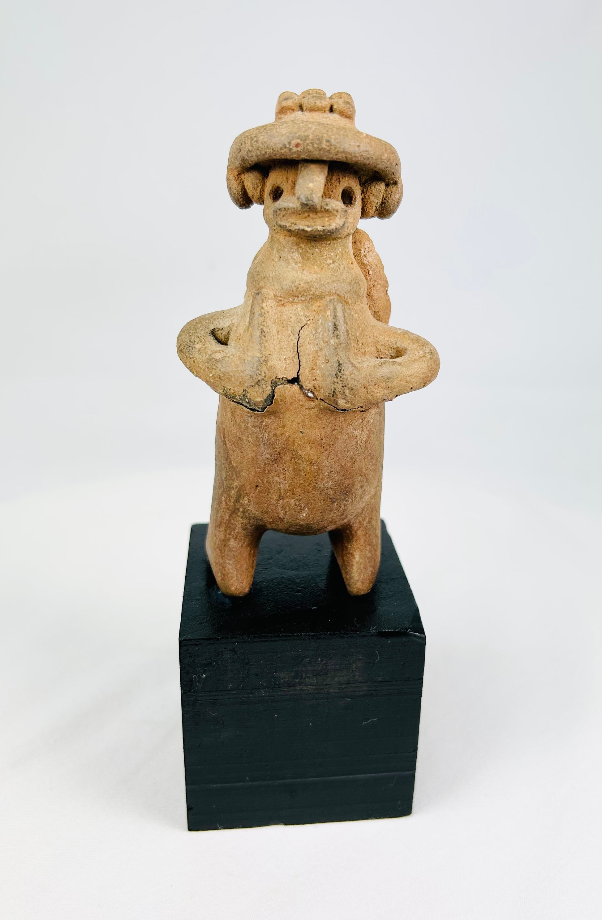 Pre-Columbian West Mexican terracotta standing effigy, likely Jalisco/Nayarit, circa 100 BCE–300 CE, hand-modeled with turban-style headdress, large earspools, hands-to-chest pose, and circular perforated back disk interpreted as warrior shield, mounted on modern display base, mineral accretions and ancient wear visible