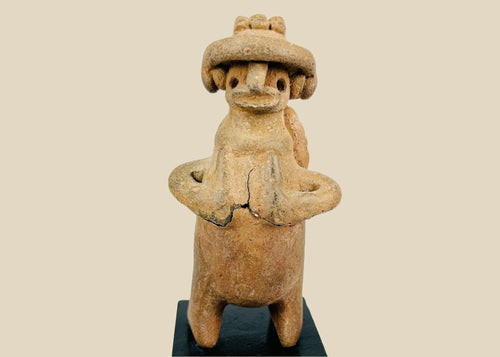Pre-Columbian West Mexican terracotta standing effigy, likely Jalisco/Nayarit, circa 100 BCE–300 CE, hand-modeled with turban-style headdress, large earspools, hands-to-chest pose, and circular perforated back disk interpreted as warrior shield, mounted on modern display base, mineral accretions and ancient wear visible