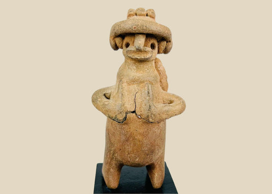 Pre-Columbian West Mexican terracotta standing effigy, likely Jalisco/Nayarit, circa 100 BCE–300 CE, hand-modeled with turban-style headdress, large earspools, hands-to-chest pose, and circular perforated back disk interpreted as warrior shield, mounted on modern display base, mineral accretions and ancient wear visible