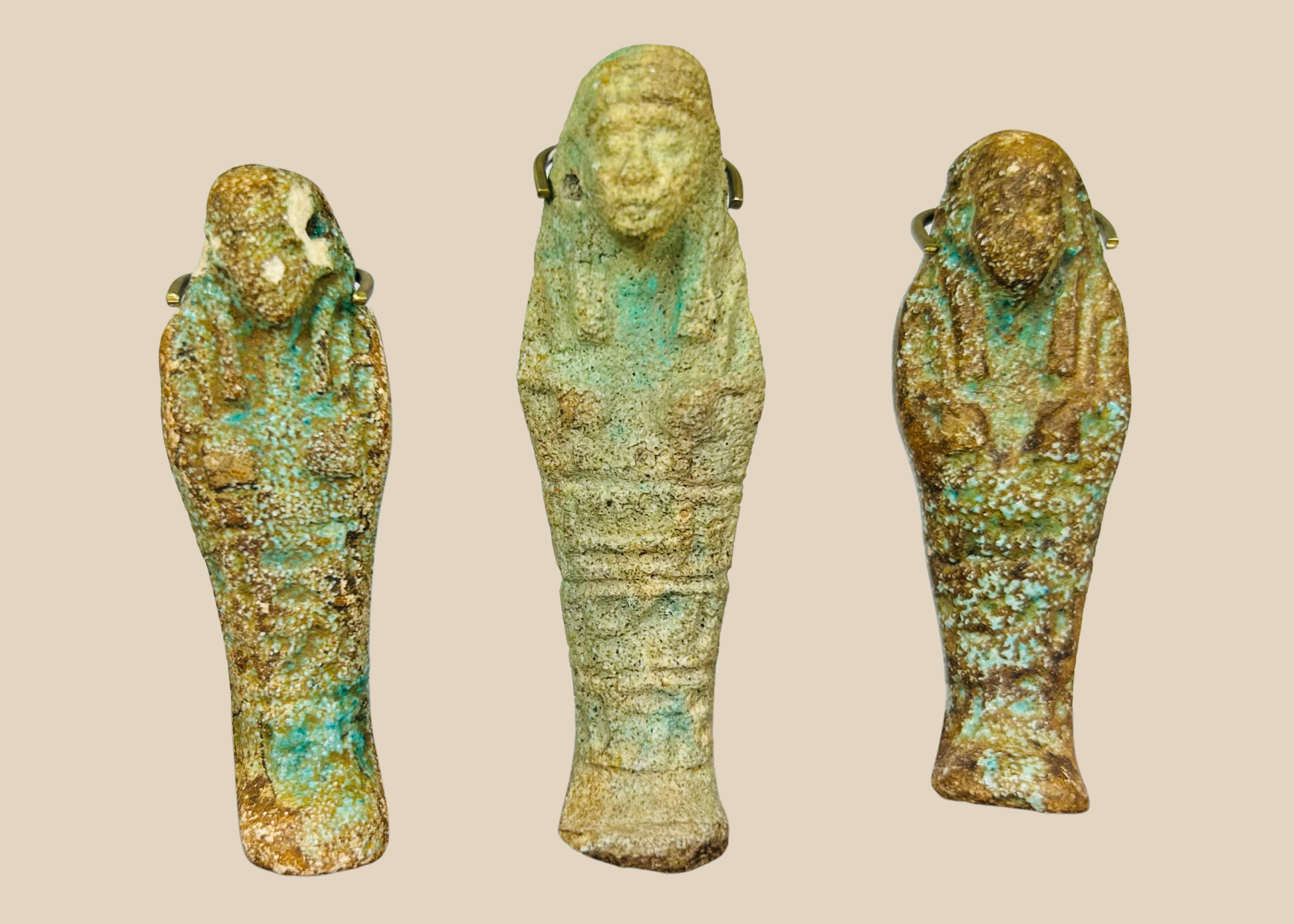 Three ancient Egyptian faience ushabti figures with turquoise and earthen patina displayed on a beige background.