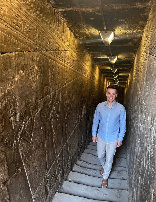 Jordan Thomas exploring an ancient Egyptian temple corridor lined with carved hieroglyphs and reliefs, reflecting Relic & Rarity's commitment to archaeological research, historical preservation, and authentic antiquities.