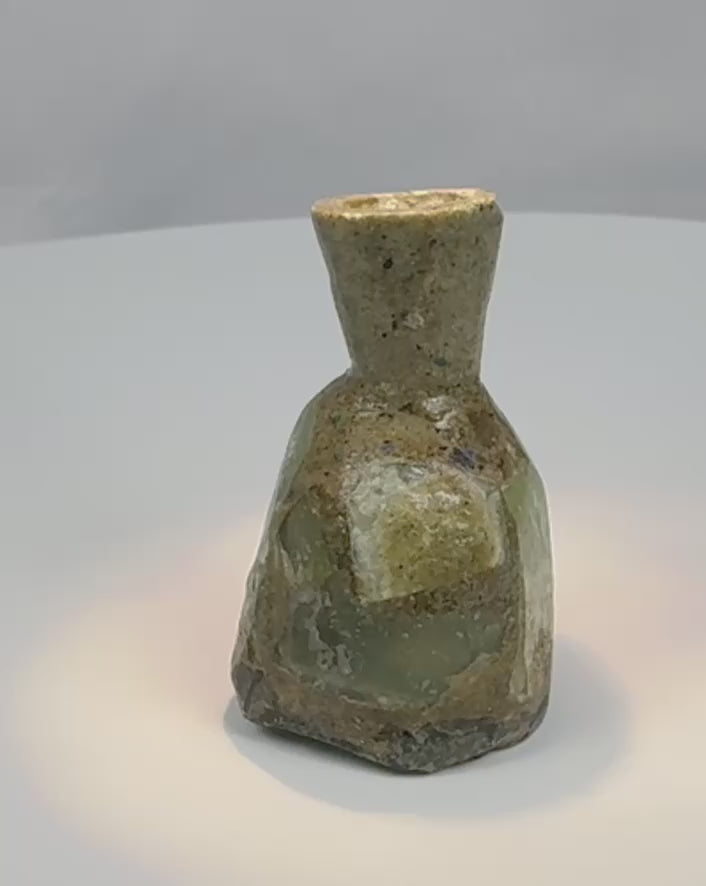 Rotating view of an ancient Roman blown green glass unguentarium with a compact body, thick cylindrical neck, narrow opening, flattened base, heavy iridescence, mineral deposits, and burial weathering