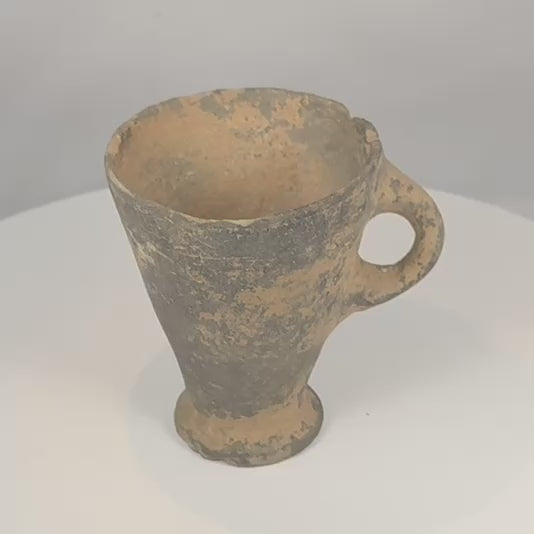 Rotating view of an ancient Near Eastern grayware pottery cup with a deep flaring conical bowl, large applied loop handle, raised circular pedestal foot, dark grey to black burnished surface, visible wheel-forming marks, pale mineral accretions, burial deposits, chipped rim, surface wear, and archaeological patina