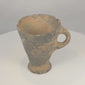 Rotating view of an ancient Near Eastern grayware pottery cup with a deep flaring conical bowl, large applied loop handle, raised circular pedestal foot, dark grey to black burnished surface, visible wheel-forming marks, pale mineral accretions, burial deposits, chipped rim, surface wear, and archaeological patina