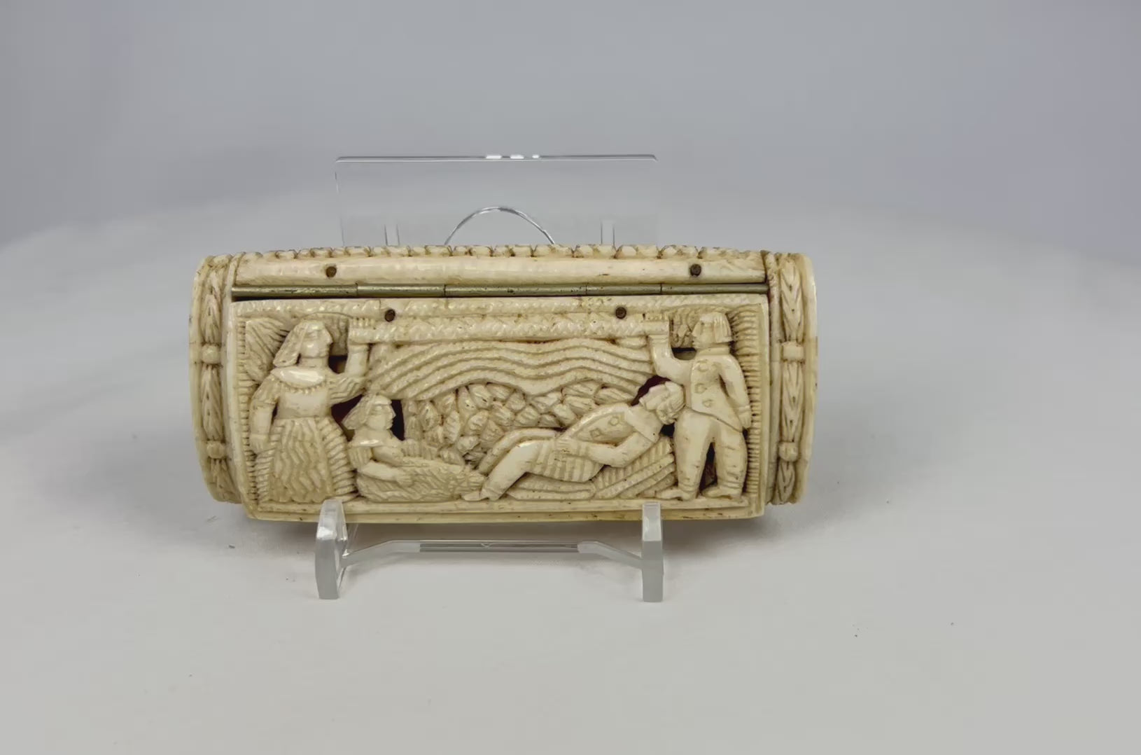 Front view of a Napoleonic POW carved bone snuff box displayed on a clear stand, showing detailed relief figures on a white background.