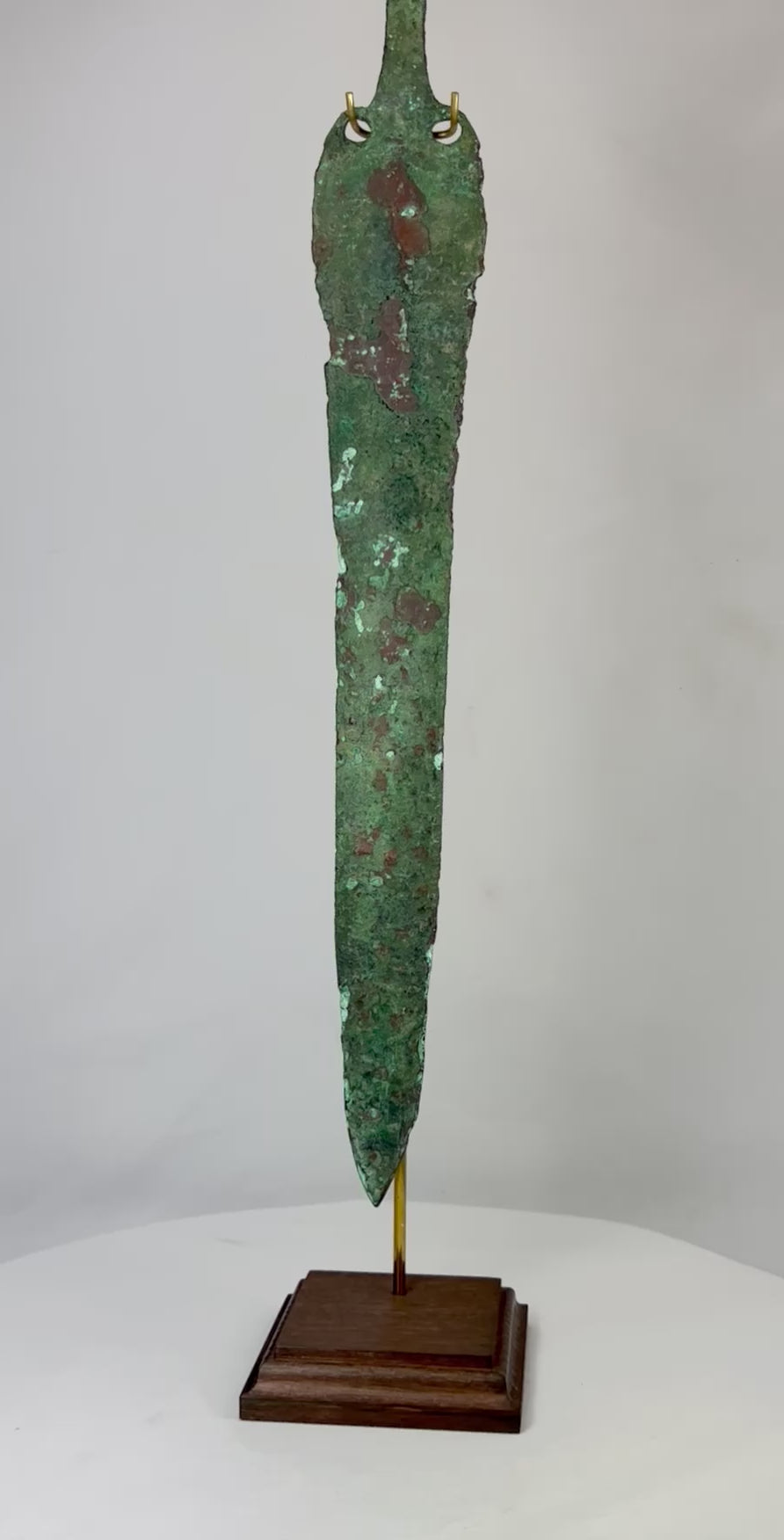 Ancient Bronze Age blade from the Caspian region, circa 2000–1000 BCE, with tapered elongated form, two perforations near the hilt, and rich green patina, mounted on a custom stand.