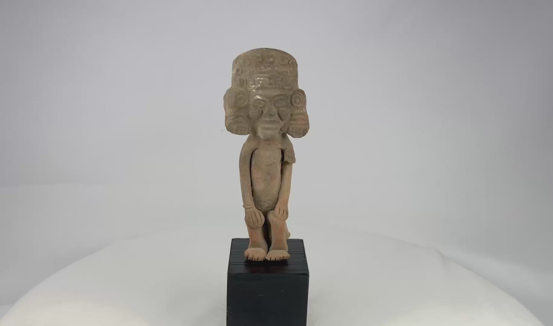 Mesoamerican Classic Period terracotta figure from Teotihuacan sphere with tall cylindrical headdress, incised roundels and step-fret band, large disk earspools, hands-on-knees posture, buff/orange clay with mineral accretions, mounted on modern black wood block, ca. 250–600 CE.
