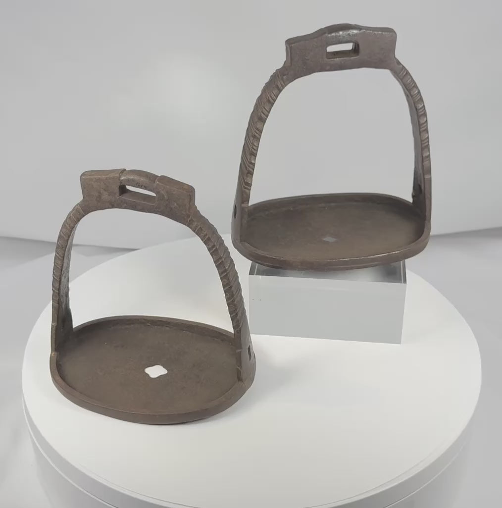Rotating view of a pair of antique Chinese hand-forged iron stirrups with broad pierced footplates, suspension loops, decorative chiseled details, and aged patina