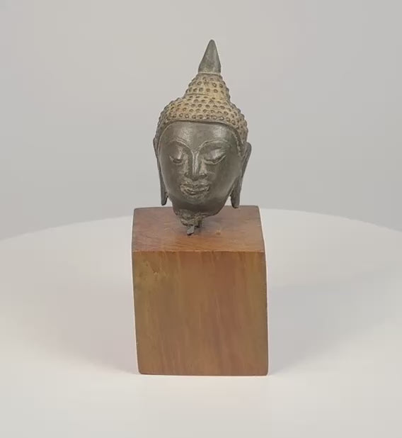 360-degree video of a Thai bronze Buddha head on a wooden base, showing textured curls, a pointed topknot, and elongated earlobes.
