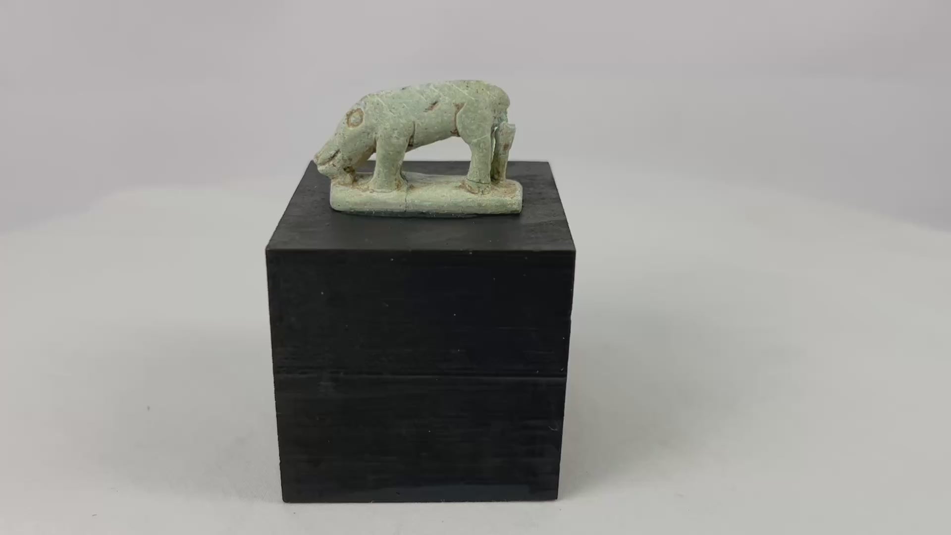 Miniature Egyptian faience shrew amulet with elongated snout, ring-and-dot eye, four legs, and rear support strut on rectangular plinth, traces of green glaze and mineral accretions, Late Period–Ptolemaic Egypt, ca. 664–30 BCE, displayed on modern wood block.