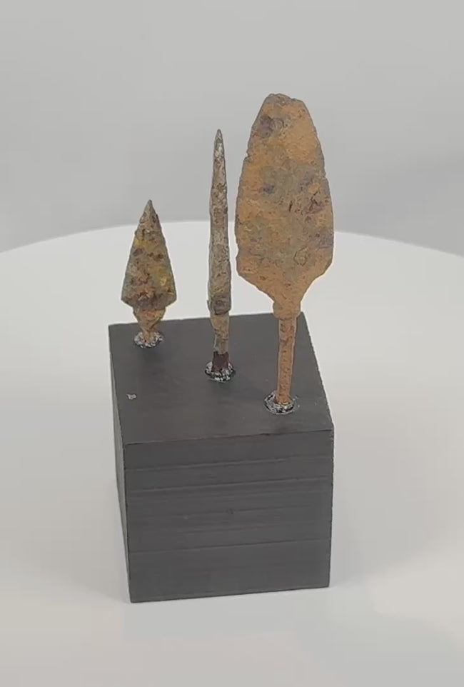 Rotating view of three ancient Roman to medieval hand-forged iron projectile points, including a broad leaf-shaped head, a narrow bodkin-style point, and a compact triangular arrowhead, each with surviving tangs, heavy oxidation, mineral encrustation, and individual black display bases