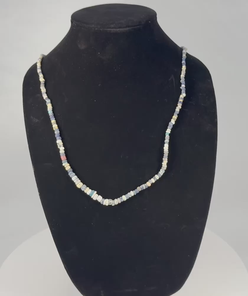 Rotating view of a restrung ancient Roman polychrome glass bead necklace with annular, cylindrical, barrel-shaped, and irregular beads in cobalt blue, pale blue, turquoise, cream, white, yellow, amber, and muted red, including striped and contrasting examples with iridescence, mineral deposits, softened edges, and archaeological weathering
