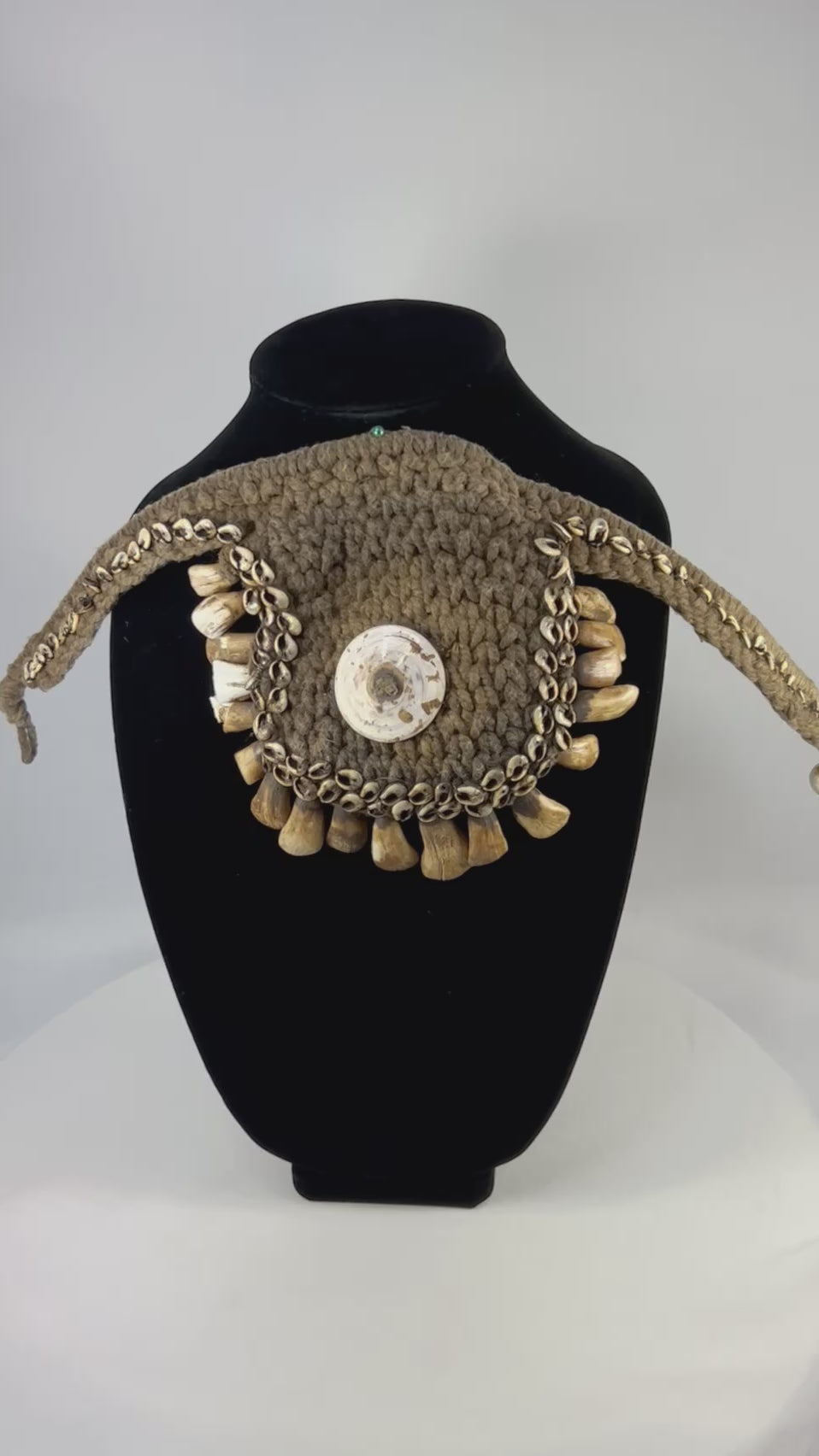Antique ceremonial adornment from Papua New Guinea, late 19th–early 20th century, woven plant fiber with central cowrie shell and fringe of pierced animal teeth, displayed on a black stand.
