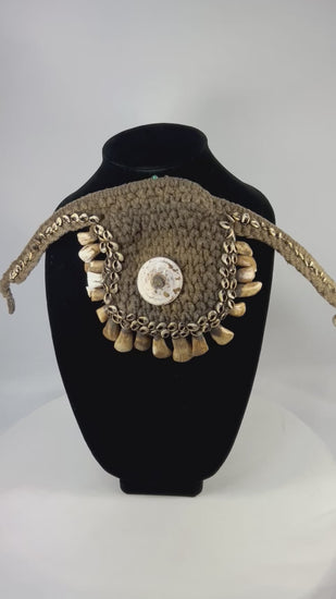 Antique ceremonial adornment from Papua New Guinea, late 19th–early 20th century, woven plant fiber with central cowrie shell and fringe of pierced animal teeth, displayed on a black stand.