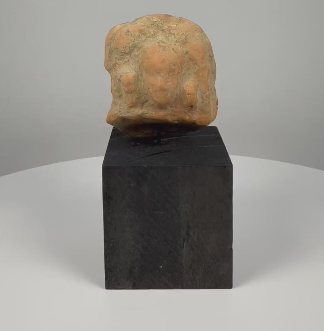 Ancient Gandharan terracotta relief plaque fragment from the 2nd–4th century CE, featuring a weathered central male bust with almond-shaped eyes, a rounded hairstyle or headdress, faint attendant figures, reddish-brown clay, natural encrustation, and a custom display base.