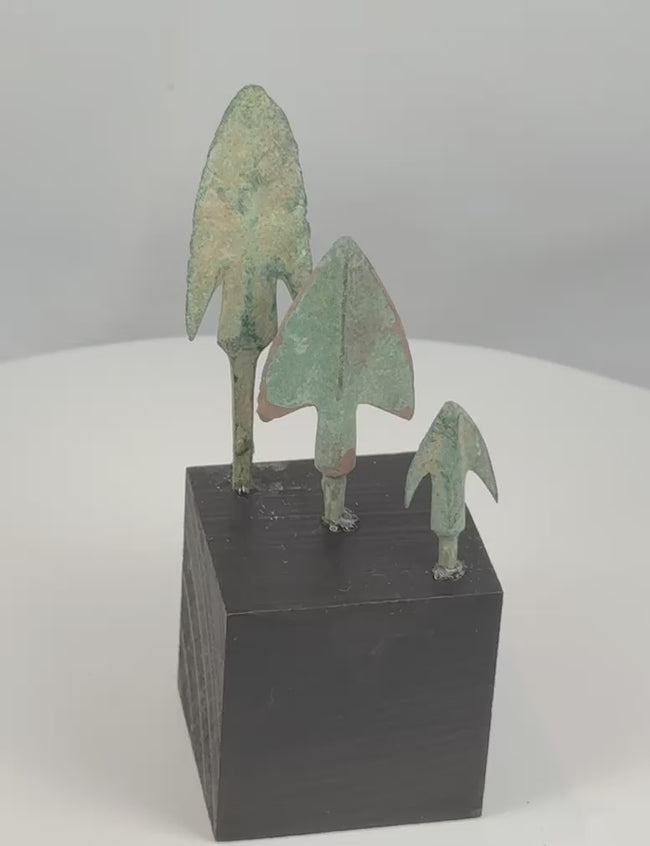 Rotating view of three ancient Western Asiatic bronze projectile points from the Late Bronze Age to Early Iron Age, including an elongated harpoon-like barbed point, a broad triangular point with a raised central rib, and a compact swallow-wing arrowhead, each with surviving tangs, green and blue-green patination, mineral encrustation, and individual black display bases