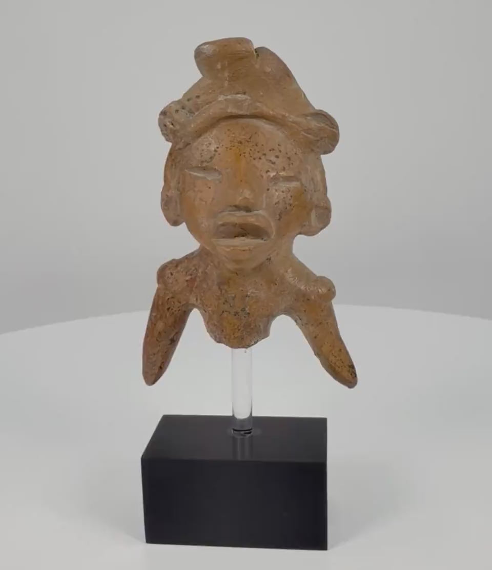 Ancient Pre-Columbian West Mexican terracotta shaft tomb figure fragment, circa 300 BCE–300 CE, featuring a sculpted headdress, incised eyes, expressive open mouth, angled arms, and weathered clay surface on a black display stand.