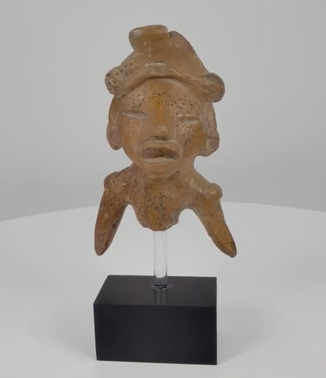 Ancient Pre-Columbian West Mexican terracotta shaft tomb figure fragment, circa 300 BCE–300 CE, featuring a sculpted headdress, incised eyes, expressive open mouth, angled arms, and weathered clay surface on a black display stand.