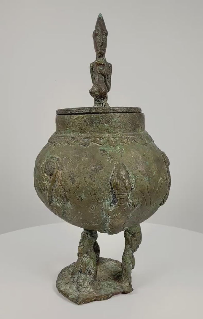 360-degree rotating video of an antique Dogon bronze ritual container from Mali, West Africa, circa 19th century, featuring a rounded vessel with engraved spiral and geometric motifs, three stylized anthropomorphic legs, and a sculpted human figure crowning the lid. The ceremonial vessel displays rich green verdigris patina, traditional Dogon metalworking craftsmanship, and age-related surface wear, shown from multiple angles.