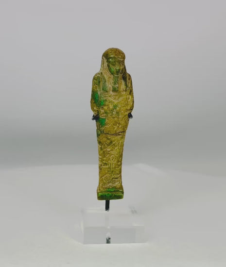 Ancient Egyptian green glazed faience Ushabti from the Late Period (664–332 BCE). Mummiform figure with tripartite wig, striated beard, and arms crossed holding hoe and pick. Hieroglyphic inscriptions adorn the body, symbolizing service in the afterlife. Reflects Egyptian funerary practices, craftsmanship, and spiritual beliefs.