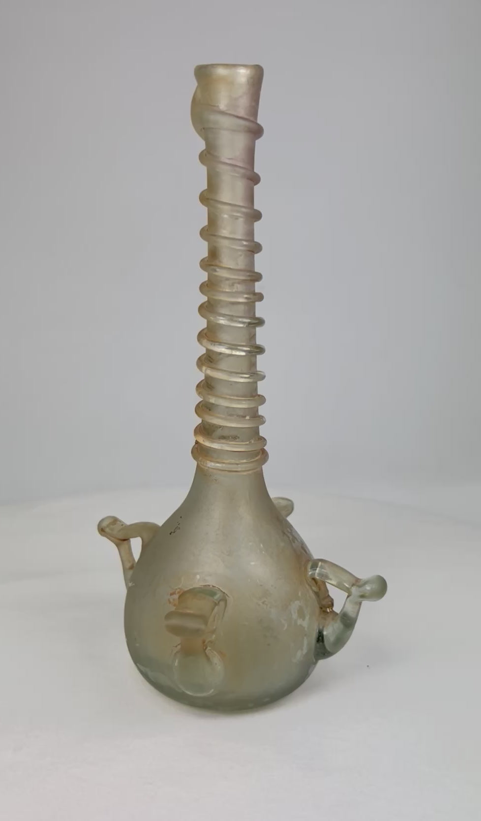 360° video of an antique Islamic blown-glass bottle featuring a spiral-threaded neck and applied handles, circa 12th–14th century CE, displayed on a neutral background.