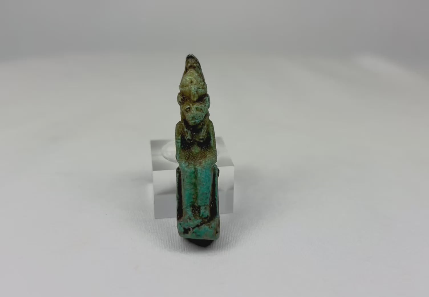 360-degree view of an ancient Egyptian seated faience deity amulet with green-blue patina on a white background.