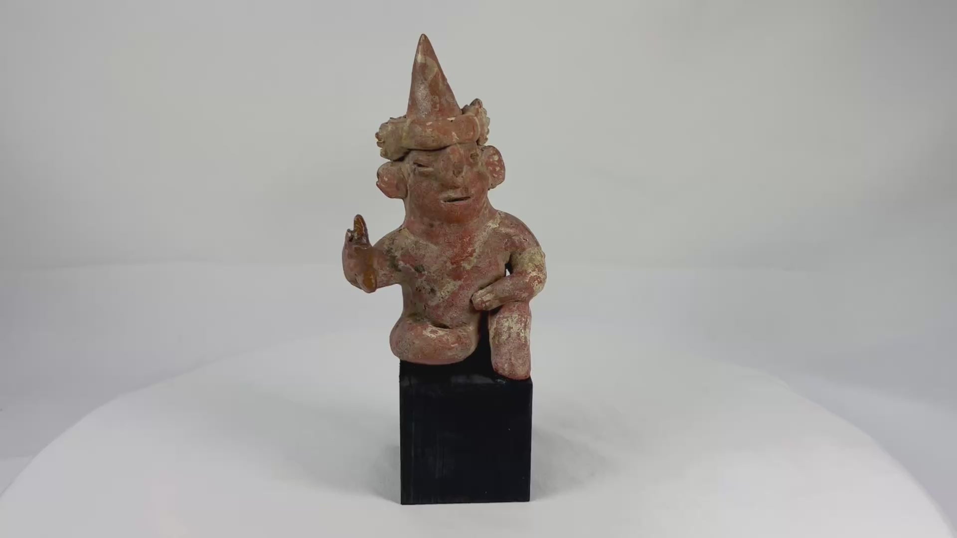 Hollow West Mexican Nayarit seated terracotta figure with tall conical headdress, turban band, large earspools, and raised right hand holding an attribute, featuring red and cream slip remnants, mineral accretions, and mounted on a modern wood display block, ca. 100 BCE–300 CE