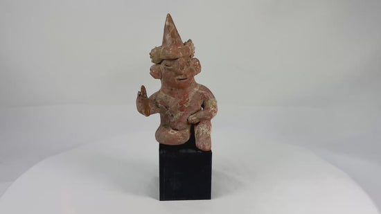 Hollow West Mexican Nayarit seated terracotta figure with tall conical headdress, turban band, large earspools, and raised right hand holding an attribute, featuring red and cream slip remnants, mineral accretions, and mounted on a modern wood display block, ca. 100 BCE–300 CE