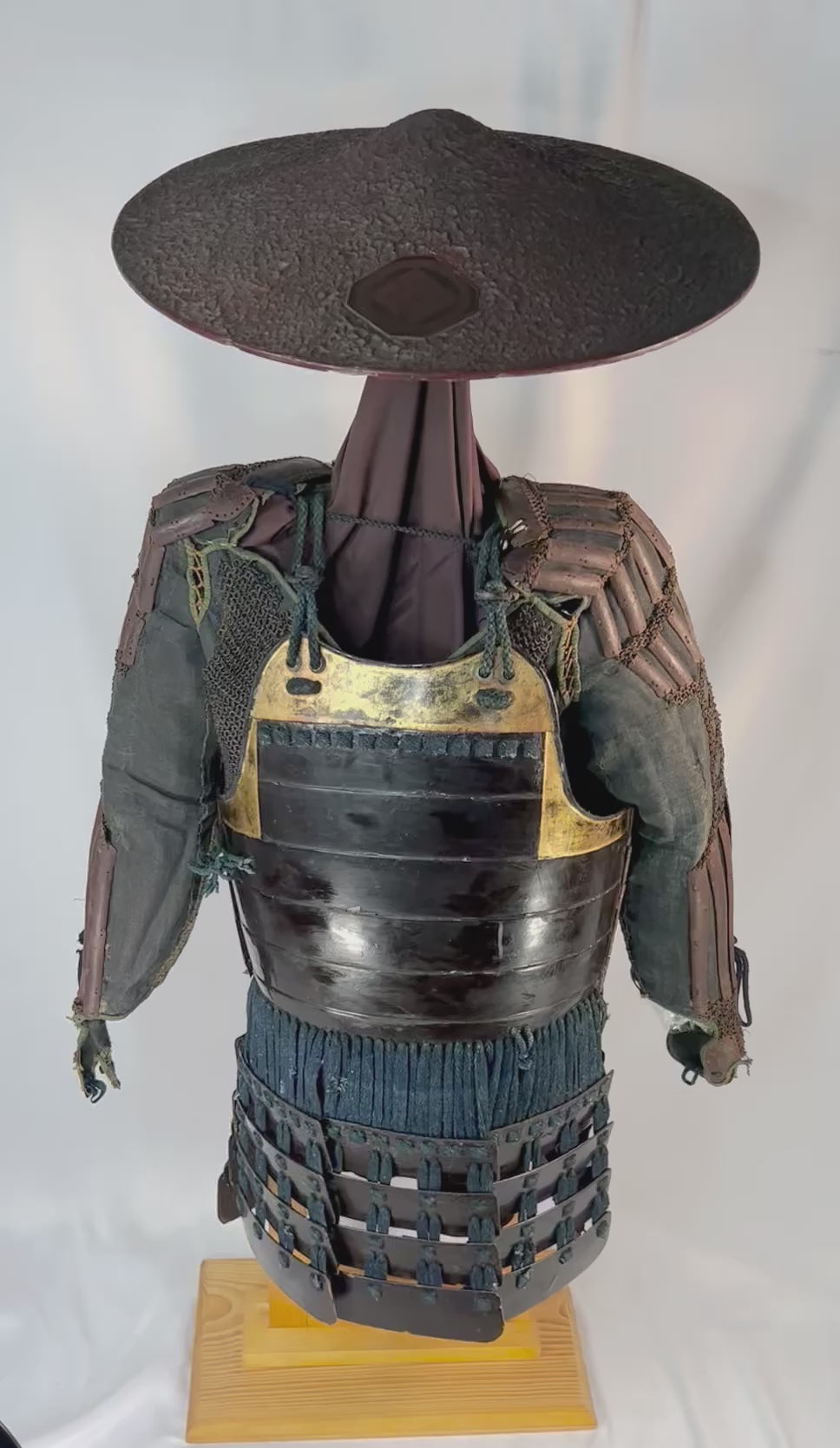 Authentic Japanese samurai armor from the Muromachi Period (1333–1573), featuring black-lacquered cuirass with gold accents, kote sleeves with chainmail reinforcement, dark-blue silk lacing, and a lacquered jingasa war hat bearing the Takeda clan crest, displayed on a wooden stand against a white background