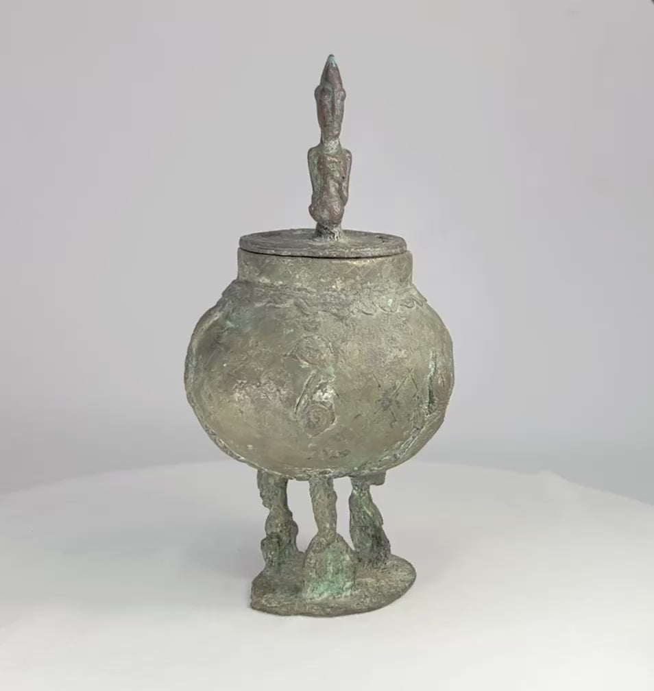 19th-century Dogon bronze container from Mali with bulbous body, engraved motifs, anthropomorphic legs, and a sculpted human figure on the lid, used in ritual and healing practices.