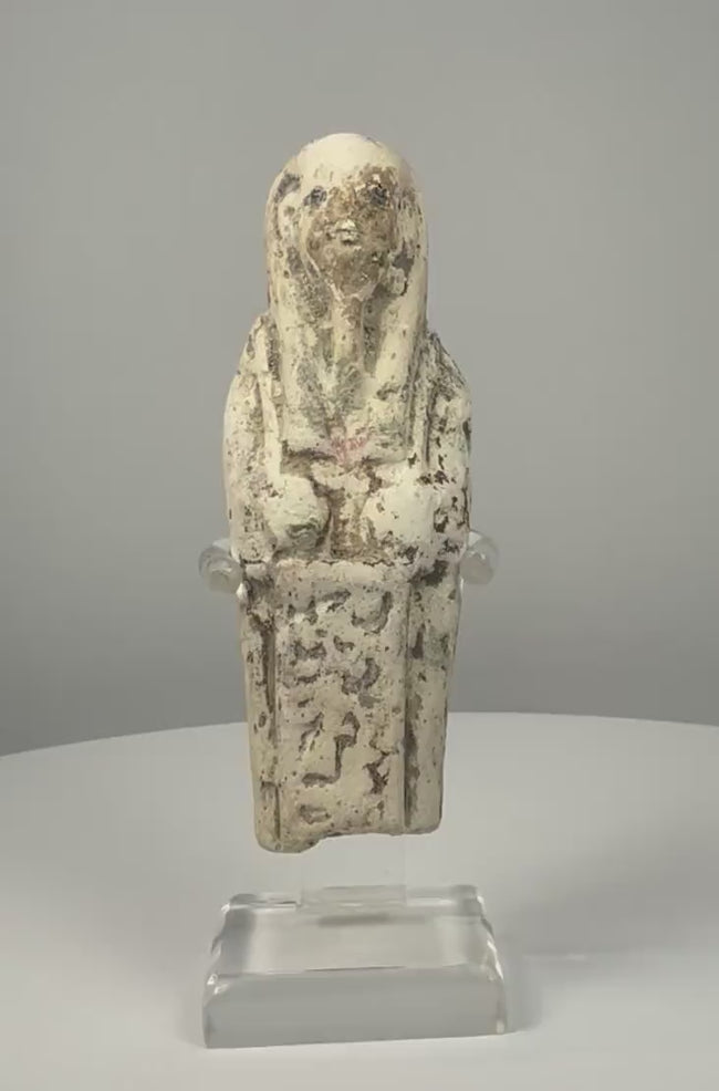360-degree view of an Egyptian limestone ushabti figure displayed on a clear acrylic stand, showing its long wig, folded arms, worn hieroglyphic inscription, and weathered cream-coloured surface.
