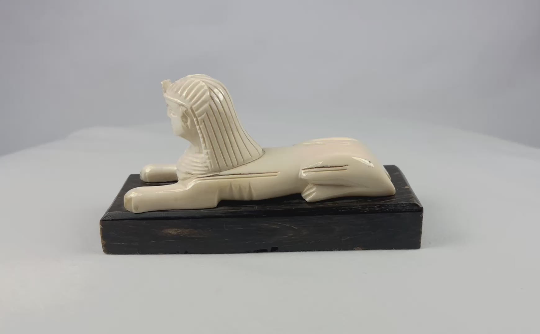 Early 20th century Egyptian revival Sphinx sculpture, reclining lion body with nemes headdress, hand-carved natural material mounted on black wooden base, decorative Egyptomania piece