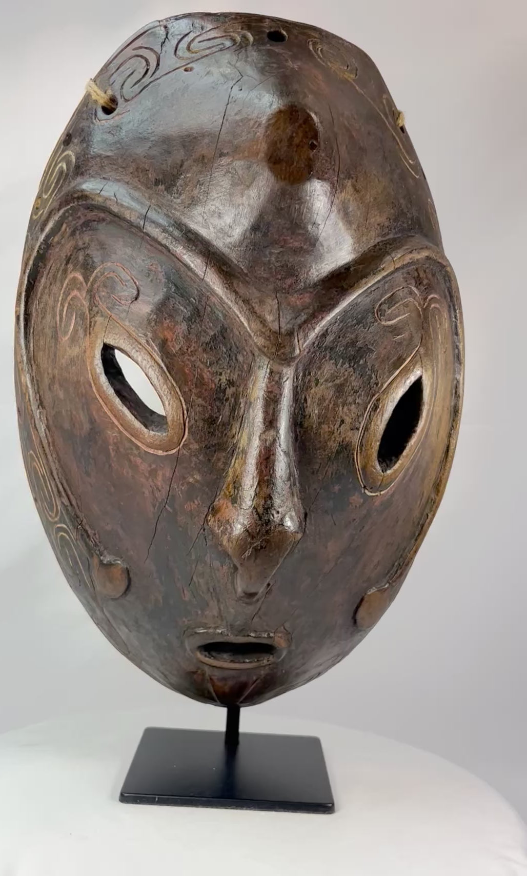Early 20th-century Sepik River tribal ceremonial mask, hand-carved from wood with elongated oval form, large almond-shaped eyes, incised swirling patterns, and traces of natural pigments, mounted on a display stand; traditional artifact used in ancestral rituals and cultural ceremonies of Papua New Guinea.
