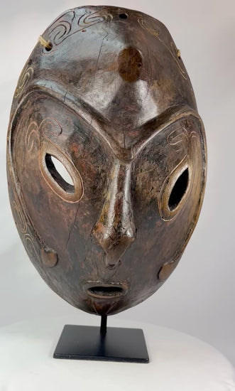 Early 20th-century Sepik River tribal ceremonial mask, hand-carved from wood with elongated oval form, large almond-shaped eyes, incised swirling patterns, and traces of natural pigments, mounted on a display stand; traditional artifact used in ancestral rituals and cultural ceremonies of Papua New Guinea.