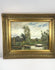 Antique early 20th-century Impressionist-style oil painting depicting a peaceful European river landscape with a solitary figure rowing a wooden boat, tall trees, lush greenery, and reflective water beneath a blue, cloud-filled sky. Presented in an ornate gilt frame with decorative foliate detailing.