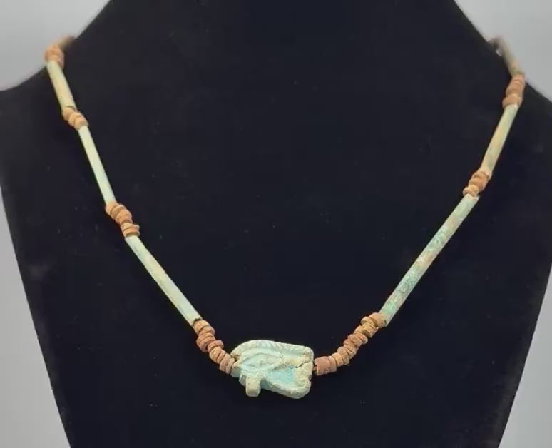 360-degree rotating view of an ancient Egyptian-style faience necklace featuring a turquoise-green Eye of Horus (Wedjat) amulet, elongated tubular beads, and earthy brown disc-shaped spacers. The video highlights the protective eye symbol, aged surfaces, intricate bead arrangement, and distinctive blue-green coloration from multiple angles.