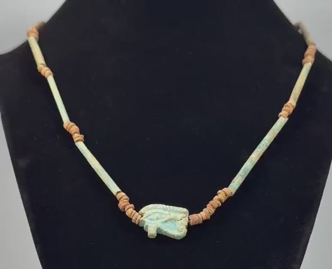 360-degree rotating view of an ancient Egyptian-style faience necklace featuring a turquoise-green Eye of Horus (Wedjat) amulet, elongated tubular beads, and earthy brown disc-shaped spacers. The video highlights the protective eye symbol, aged surfaces, intricate bead arrangement, and distinctive blue-green coloration from multiple angles.