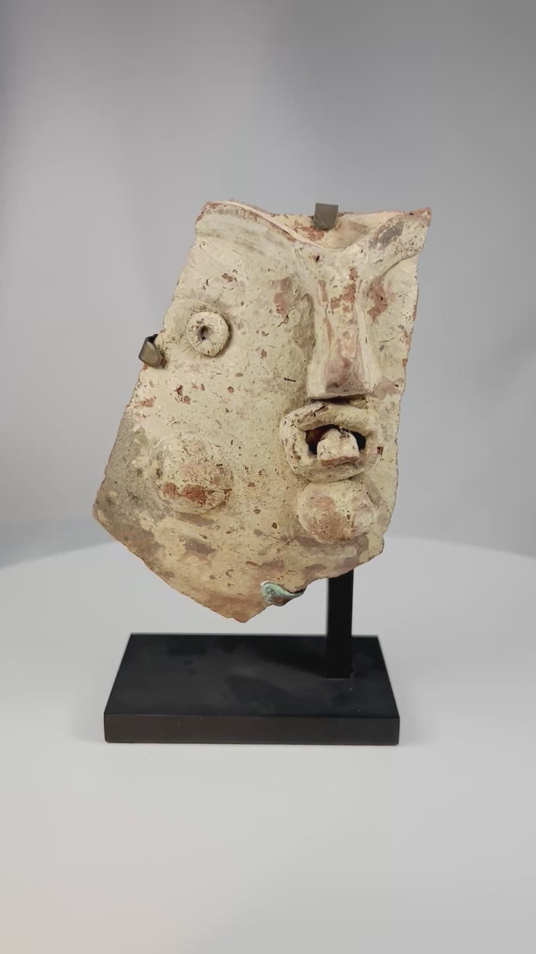 360-degree view of an ancient Greco-Roman terracotta theatrical mask fragment from the Hellenistic to Roman period, mounted on a display stand showing sculpted facial features and expressive dramatic styling.
