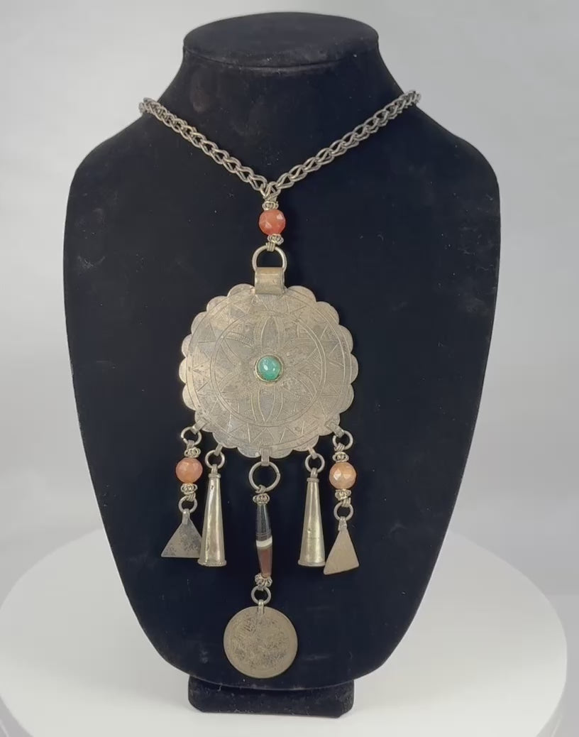 Rotating view of a mid-20th-century Central Asian tribal necklace with a large scalloped circular pendant, engraved six-petalled rosette, turquoise-coloured central cabochon, faceted orange beads, banded bead, conical and triangular metal drops, worn coin with Arabic or Persian script, interlinked metal chain, handmade suspension rings, tarnishing, oxidation, and aged patina