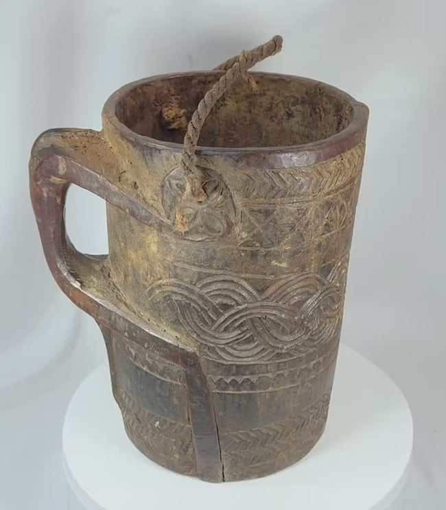 360-degree view of an antique Himalayan carved wooden storage vessel featuring geometric relief carving, an integrated side handle, original fiber rope suspension handle, and rich aged patina.