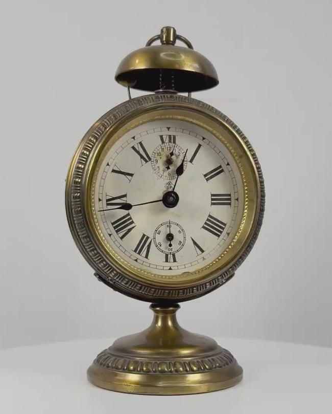 360-degree rotating view of an antique German JUF brass pedestal alarm clock, circa 1900–1920, featuring an ornate circular brass case, Roman numeral dial, subsidiary registers, domed alarm bell, decorative pedestal base, and rear mechanical winding controls.