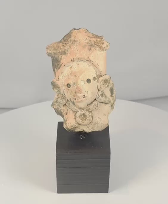 Rotating view of a Pre-Columbian terracotta figural fragment with a rounded face, perforated eyes, elaborate headdress, circular earspools, neck ornaments, and archaeological surface deposits