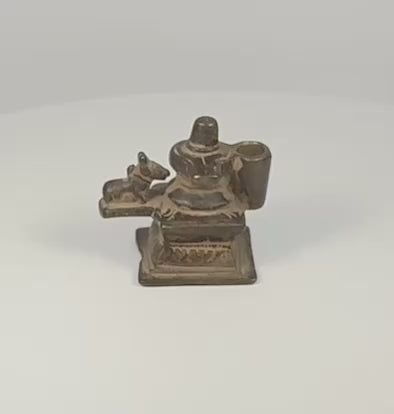 360-degree rotating view of a 19th century Indian brass Shiva Lingam and Nandi miniature devotional shrine, showing the stepped pedestal, recumbent Nandi bull, cylindrical fitting, and aged dark patina.