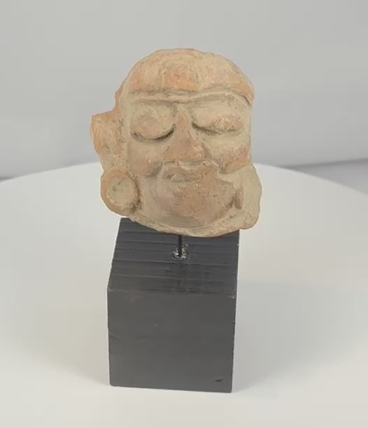 Rotating view of an ancient Mesoamerican terracotta head fragment with heavily lidded eyes, circular ear ornaments, pigment traces, and a weathered archaeological surface