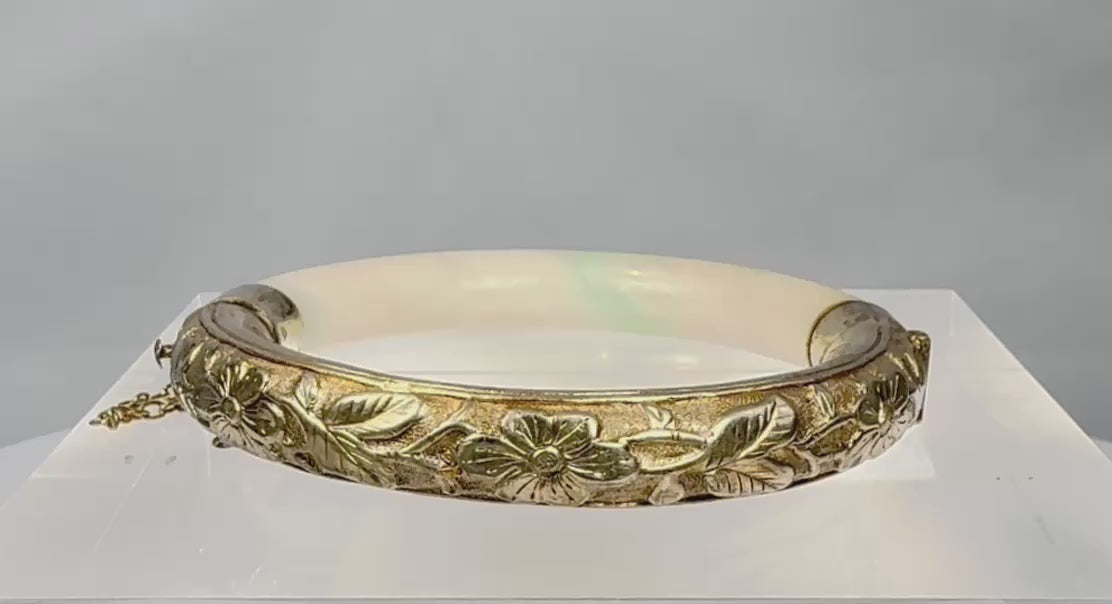 Rotating view of an antique Chinese hinged jade or hardstone bracelet with pale celadon colouring, natural green inclusions, repoussé floral silver mounts, a safety chain, and an interior SILVER mark