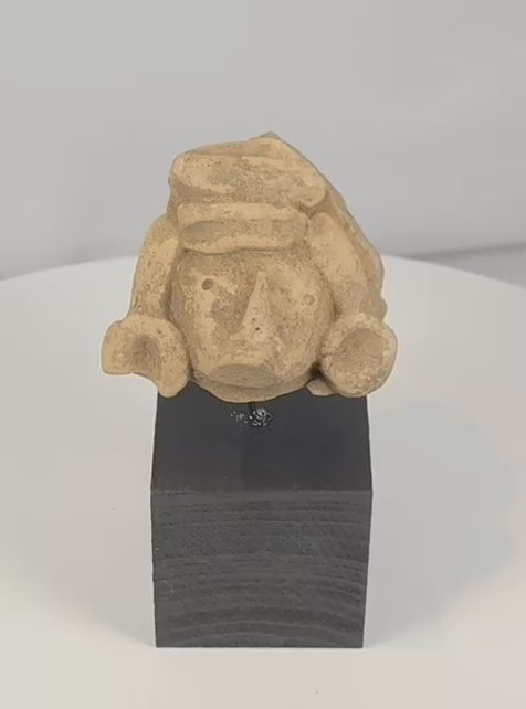 Rotating view of a Pre-Columbian carved stone head fragment with recessed eyes, a triangular nose, circular earspools, an elaborate headdress, weathered surfaces, and a custom display stand