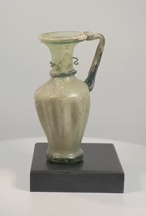 360-degree rotating view of an ancient Roman–Byzantine glass jug dating to the 4th–5th century CE, featuring pale green glass, vertical ribbing, a flared mouth, applied blue-green glass decoration, a curved handle, and extensive ancient surface patination.